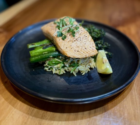 Oven-baked salmon fillet served over herb rice with broccolini, asparagus, microgreens, and a lime wedge on a black plate.