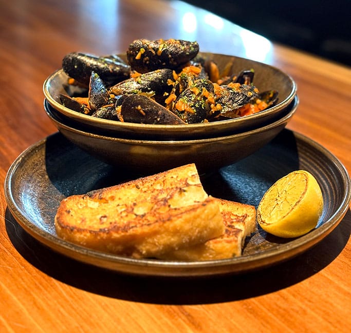 PEI-Mussels-mobile Bowl of PEI mussels in a rich garlic herb sauce, served with toasted grilled bread and a charred lemon wedge on a dark plate atop a wooden table.