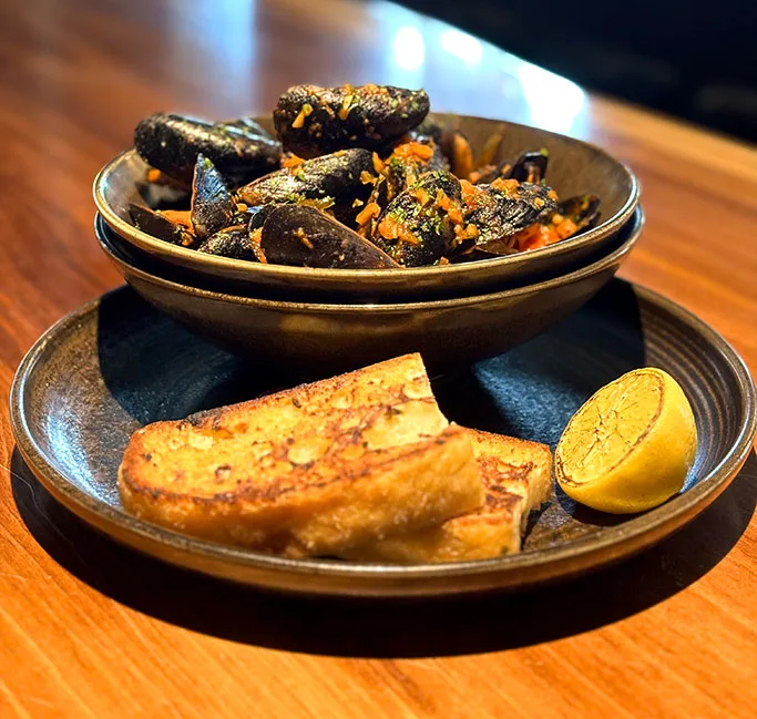 PEI-Mussels-mobile Bowl of PEI mussels in a rich garlic herb sauce, served with toasted grilled bread and a charred lemon wedge on a dark plate atop a wooden table.