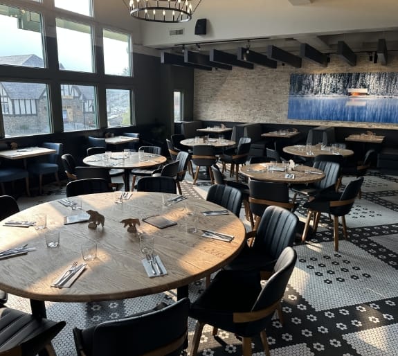 Spacious main dining room at The Boss Kitchen & Bar in Banff featuring round wooden tables, dark upholstered chairs, decorative tiled flooring, and large windows.