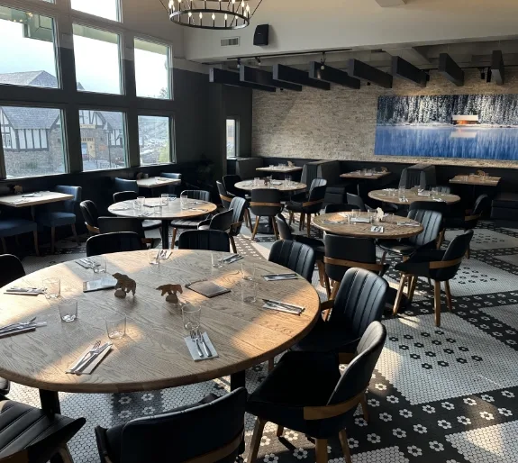 the-boss-banff-main-dining-room-tables-mobile Spacious main dining room at The Boss Kitchen & Bar in Banff featuring round wooden tables, dark upholstered chairs, decorative tiled flooring, and large windows.